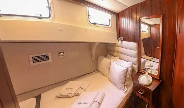 A cozy cabin aboard Ulucinar with a bed, cushions, wood panels, and small porthole window