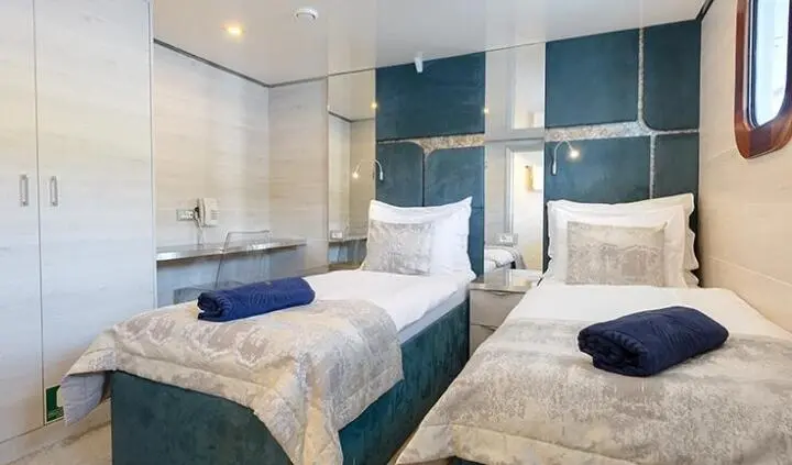 Twin cabin on yacht Yolo with two beds, blue accents, seating area, and ensuite window view