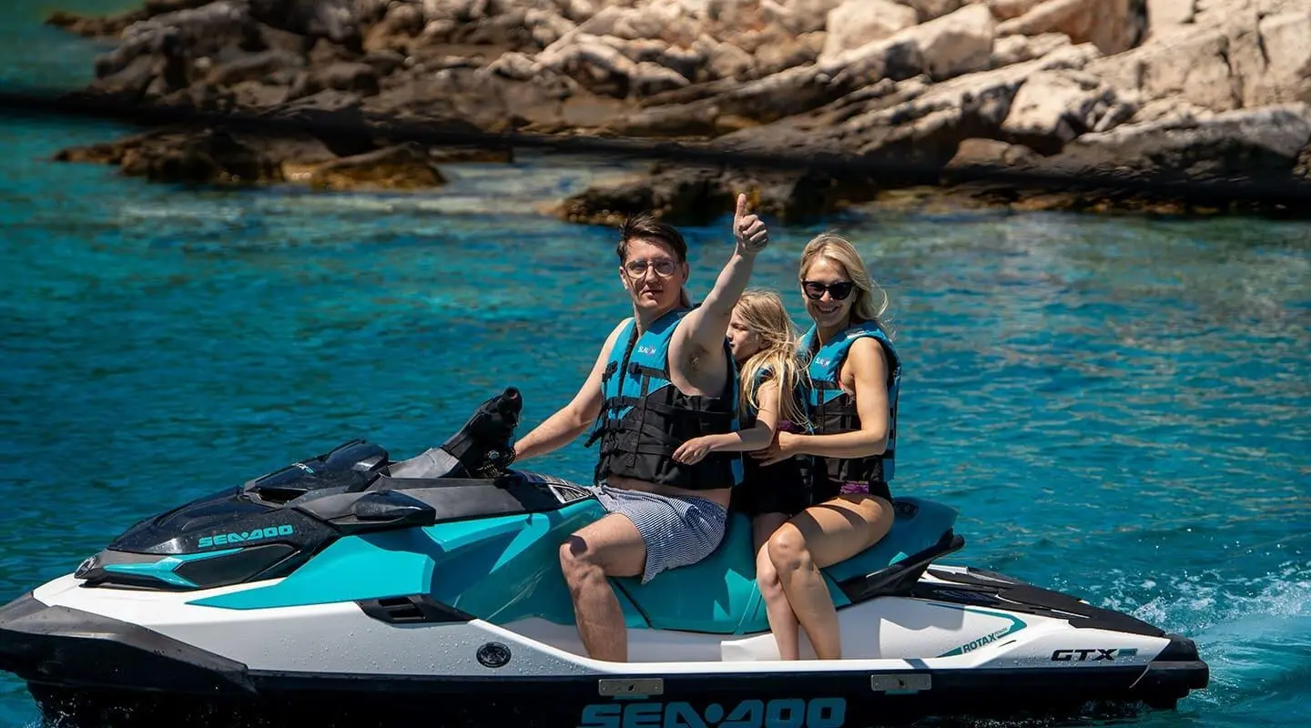 Two people sit on a sleek yacht bow near rocky coastline and clear blue sea