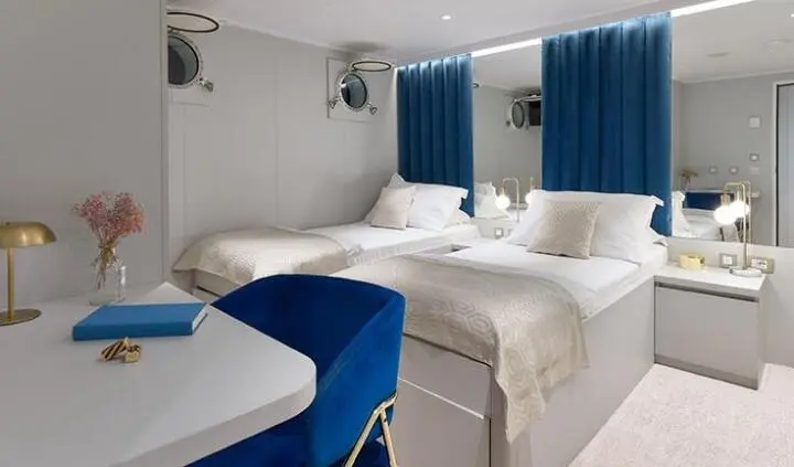 Bellezza cabin interior with twin beds, white linens, blue accents, and curtains