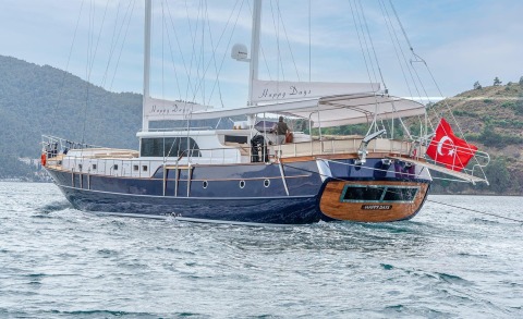 Luxurious blue-hulled yacht with 'Happy Days' canopy on deck, Turkish flag at stern, anchored near a hilly coastline on the open sea.