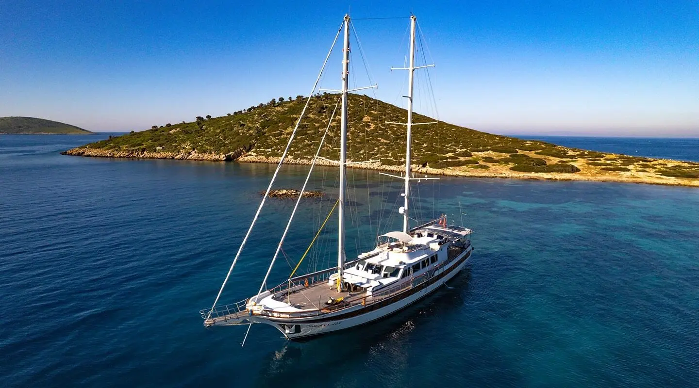 Two-masted sailing yacht anchored in a calm cove with a rocky island shore nearby