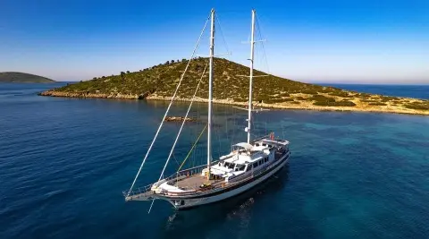 Two-masted sailing yacht anchored in a calm cove with a rocky island shore nearby