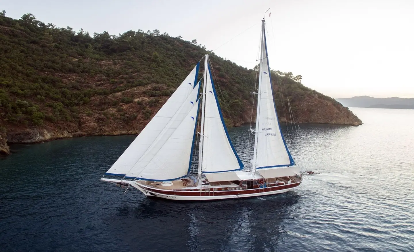 Kayhan Kaptan yacht with two white-sailed mizzen boats near rocky coastline at sunset