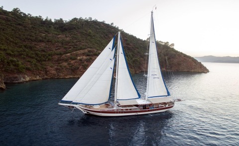 Kayhan Kaptan sailing yacht near rocky coastline with white sails and blue trim