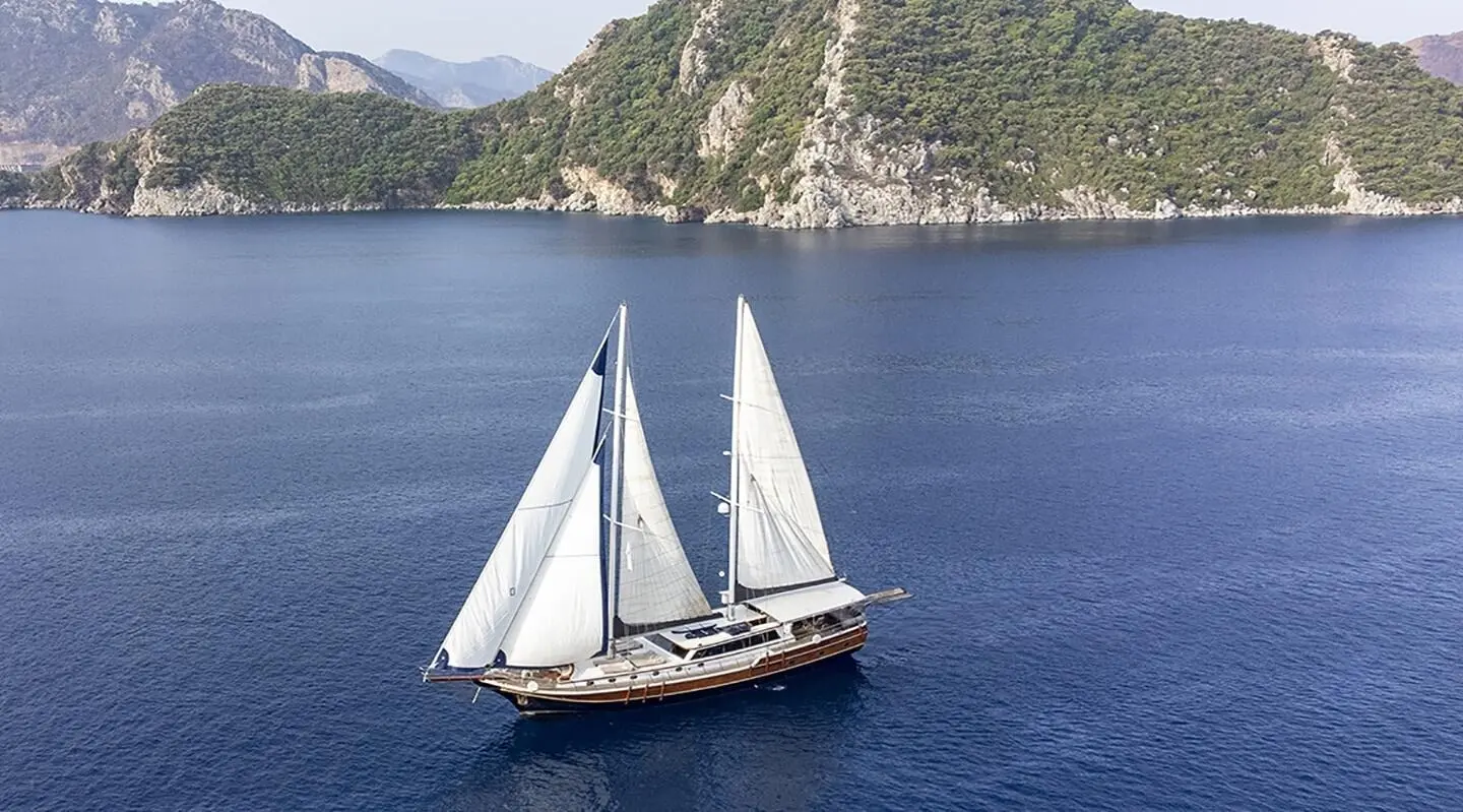 Diva Deniz yacht with white sails gliding on blue sea near rocky coastline at sunset