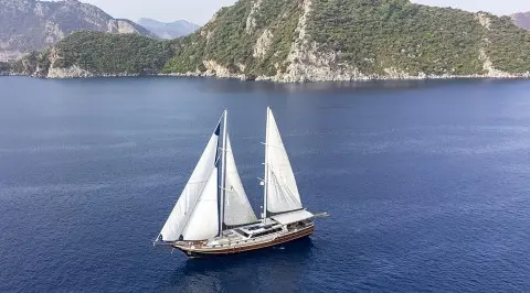Diva Deniz yacht with white sails gliding on blue sea near rocky coastline at sunset