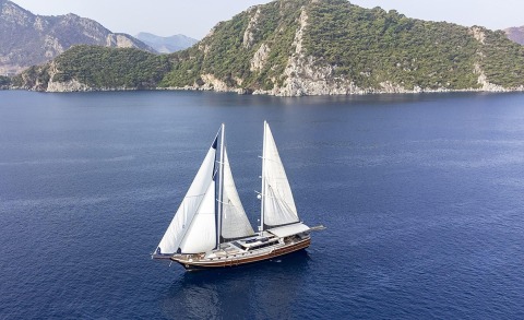 Diva Deniz gulet yacht with two white sails on blue sea, sails up near rocky green hills in the background