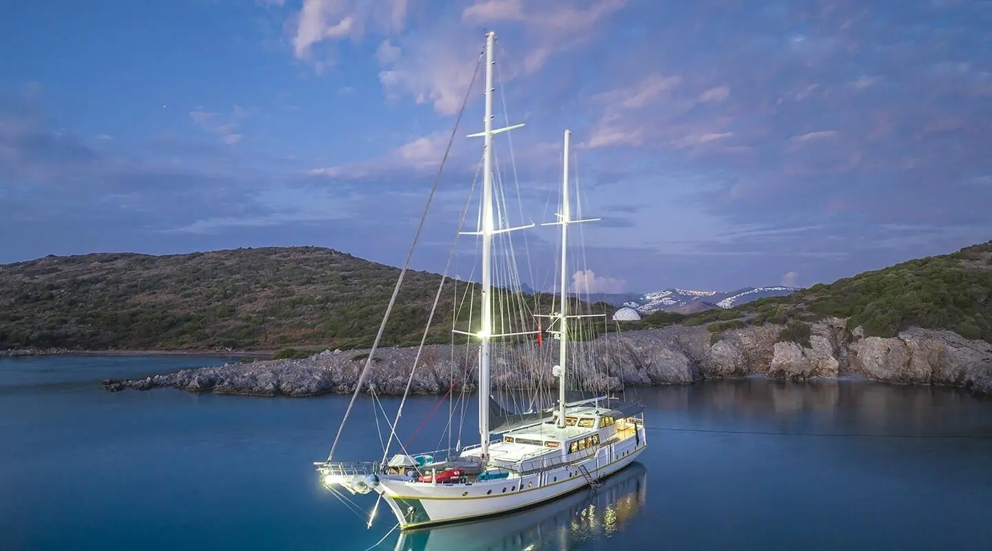 A green and white sailboat anchored in calm blue waters near rocky coast at sunset