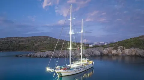 A green and white sailboat anchored in calm blue waters near rocky coast at sunset