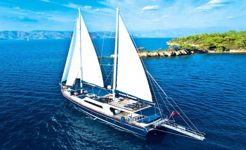 Two-masted sailing yacht Maske 2 sails on blue sea, with coastline and green trees in the background