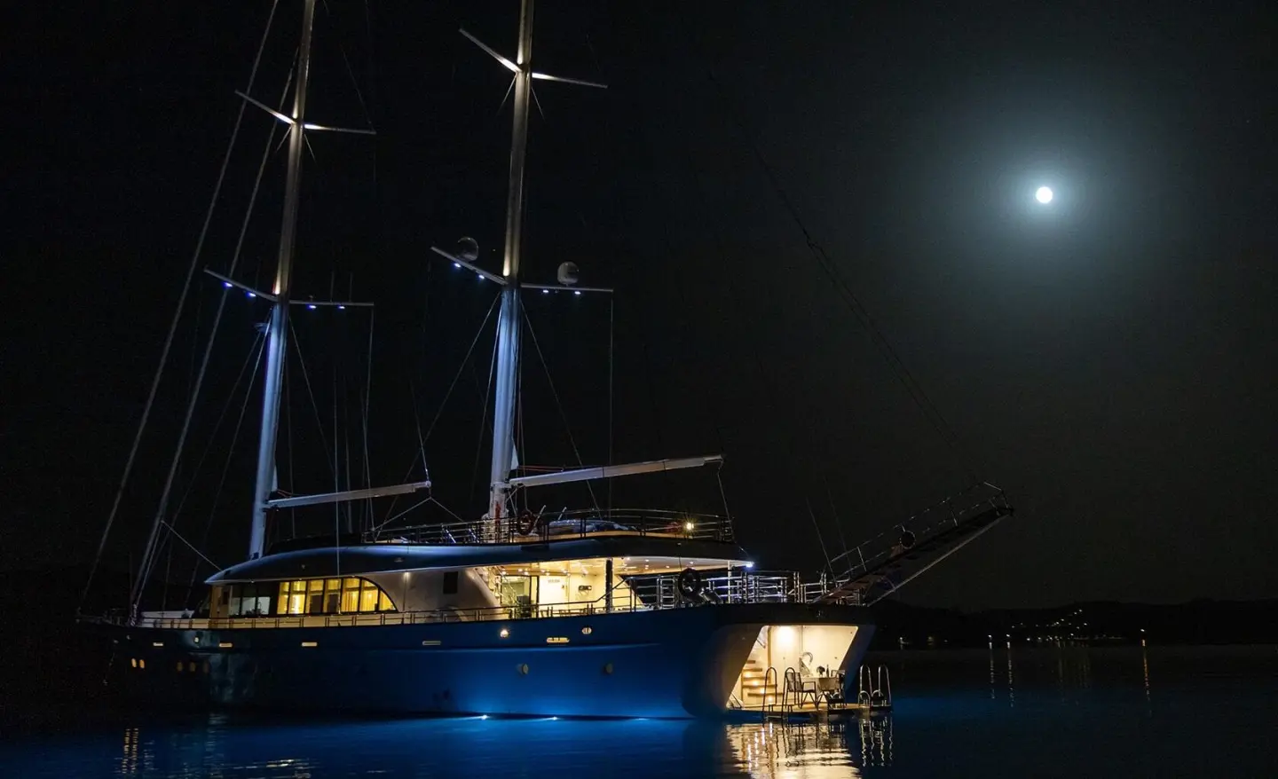 Two-masted yacht at night with blue lights and moon reflected on water