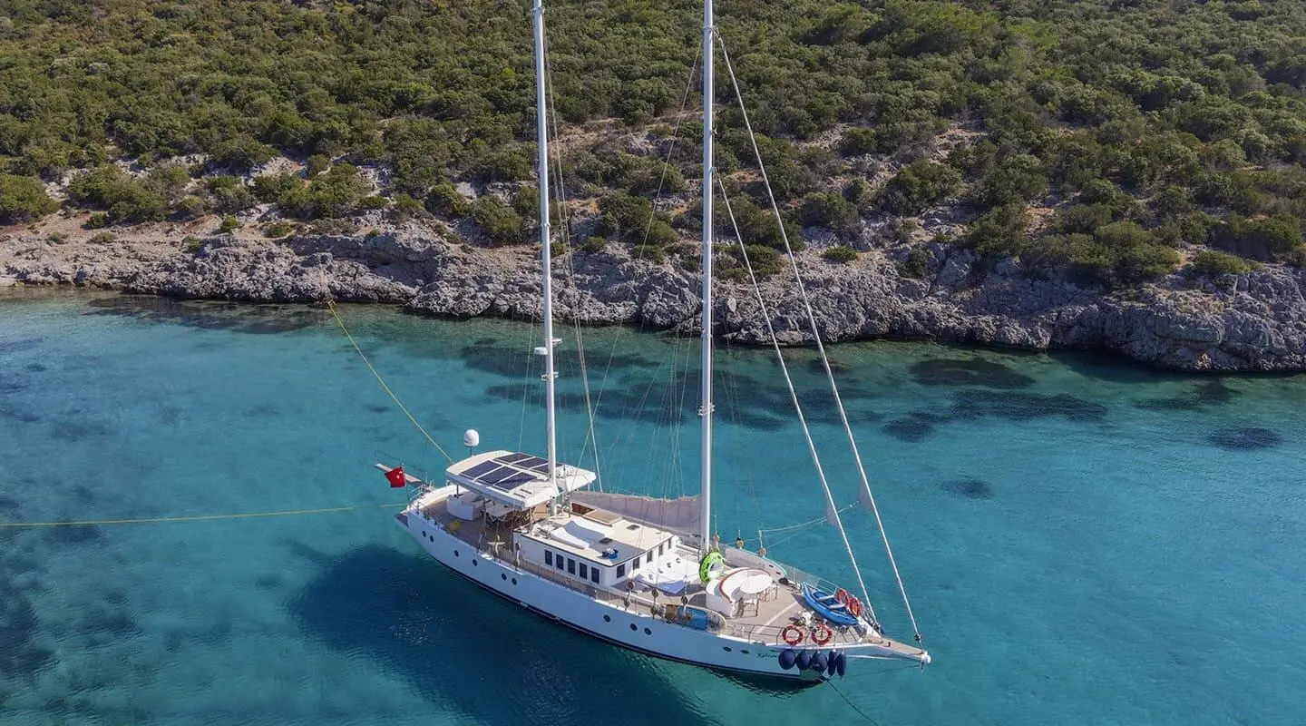 Two-masted luxury sailing yacht anchored near rocky coastline with turquoise water