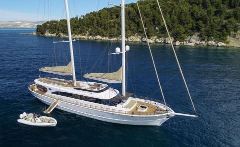 Acapella motor-sail yacht anchored in blue water near rocky green shoreline