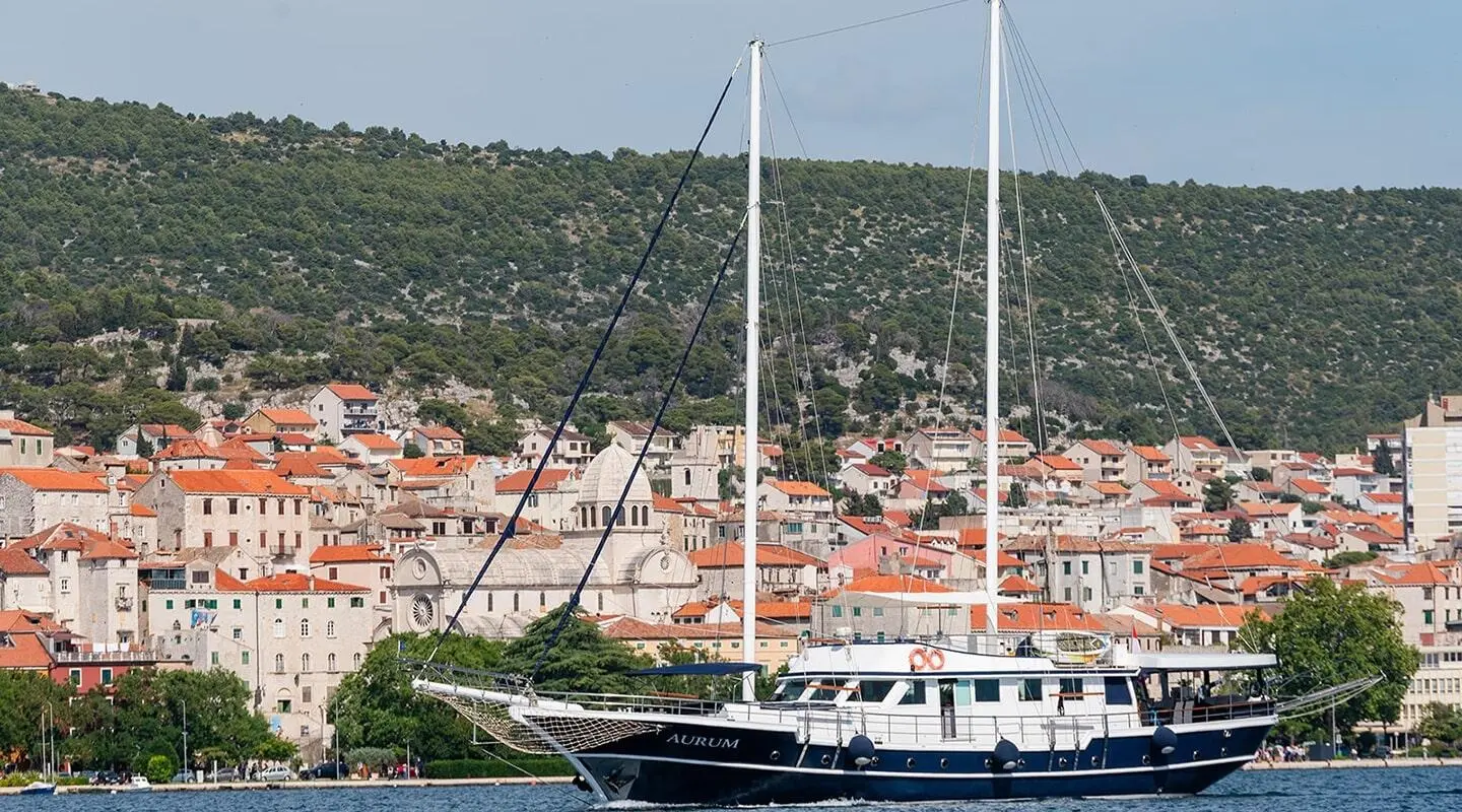 Auriun? No, image shows a luxurious sailing yacht named Aurum docked by a coastal town with red-tiled roofs at sunset