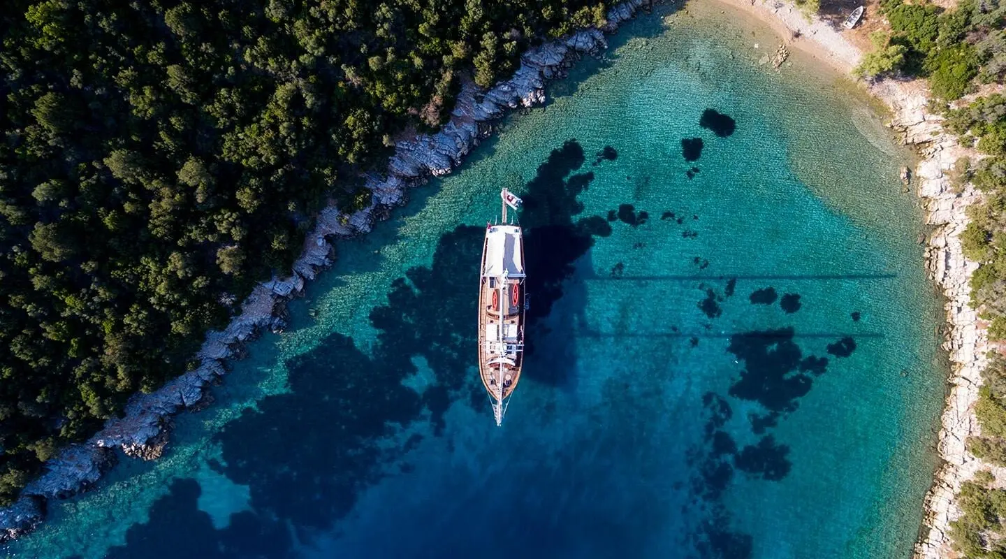 Aerial view of a white gulet anchored in turquoise clear water near a rocky shore