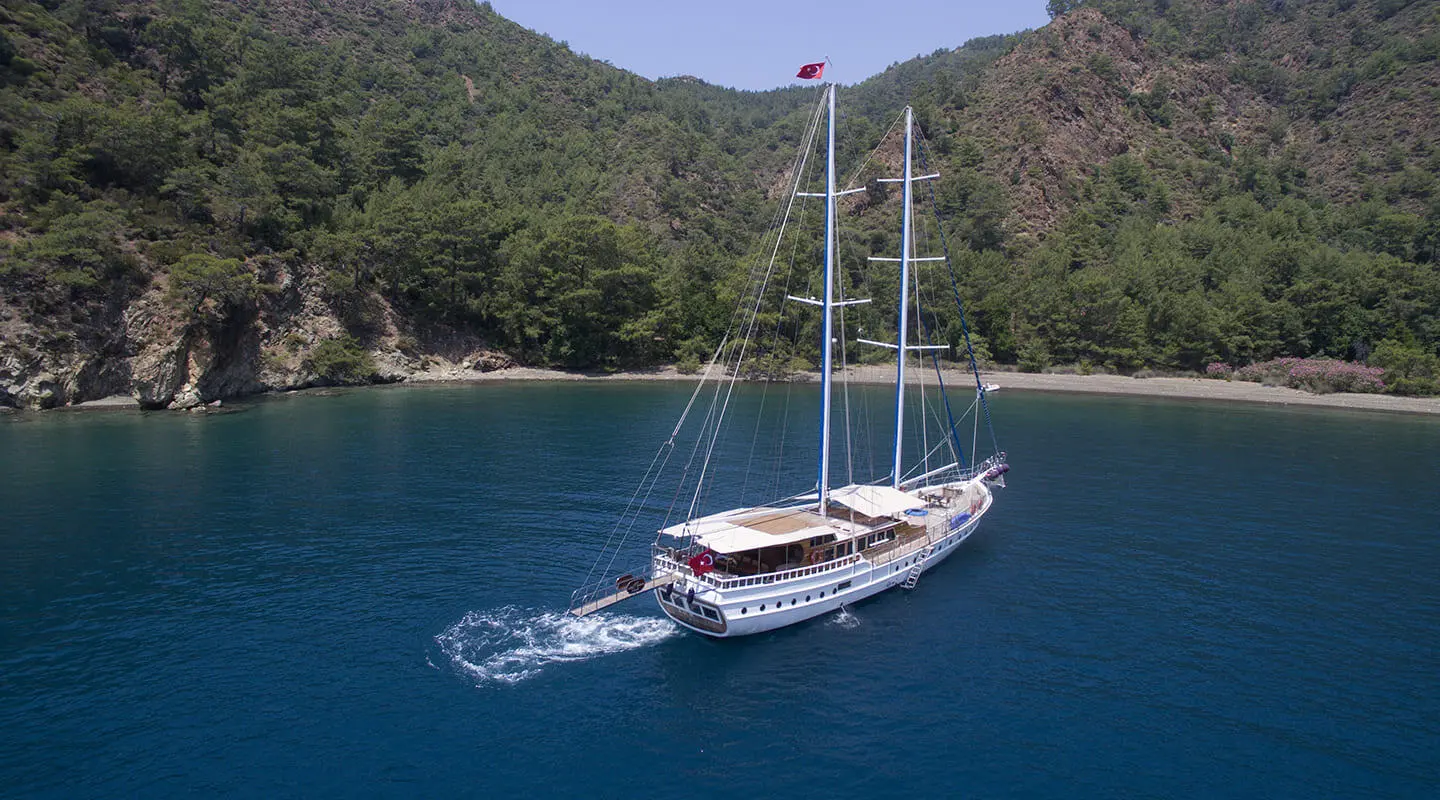 White two-masted gulet yacht anchored in a blue bay with forested coastline