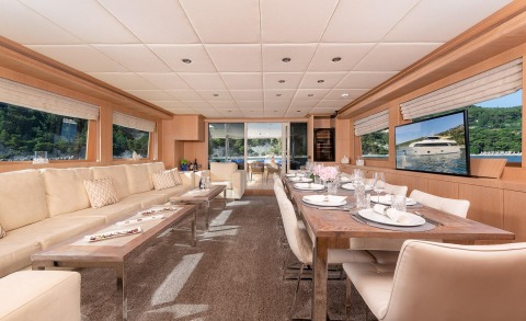 Interior of Grace luxury motor yacht with long wooden table set for dining, beige upholstered seating, plates and flowers, visible doorway to deck and water view outside