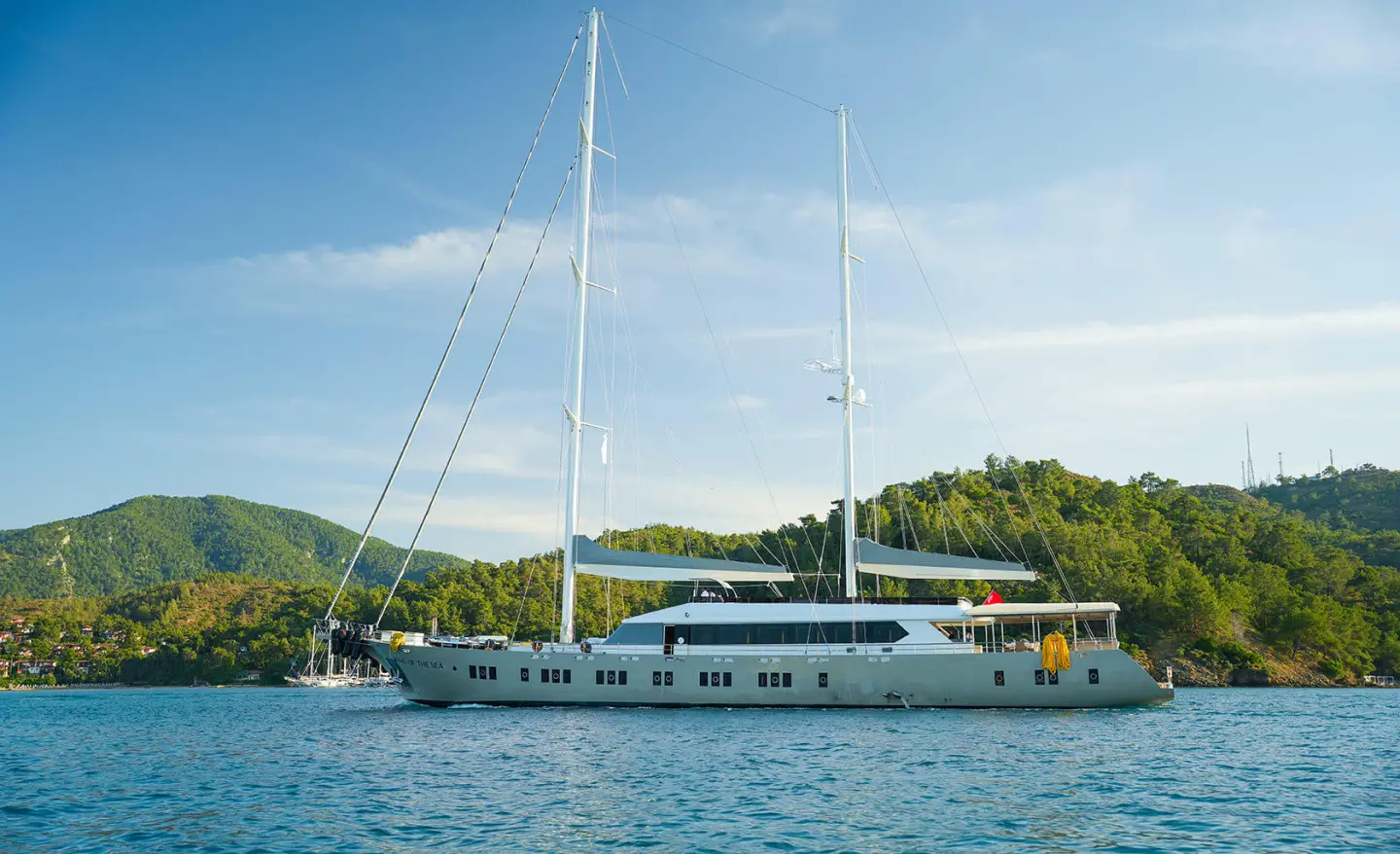 Two-masted yacht anchored by the shore with green hills and blue sky in the background