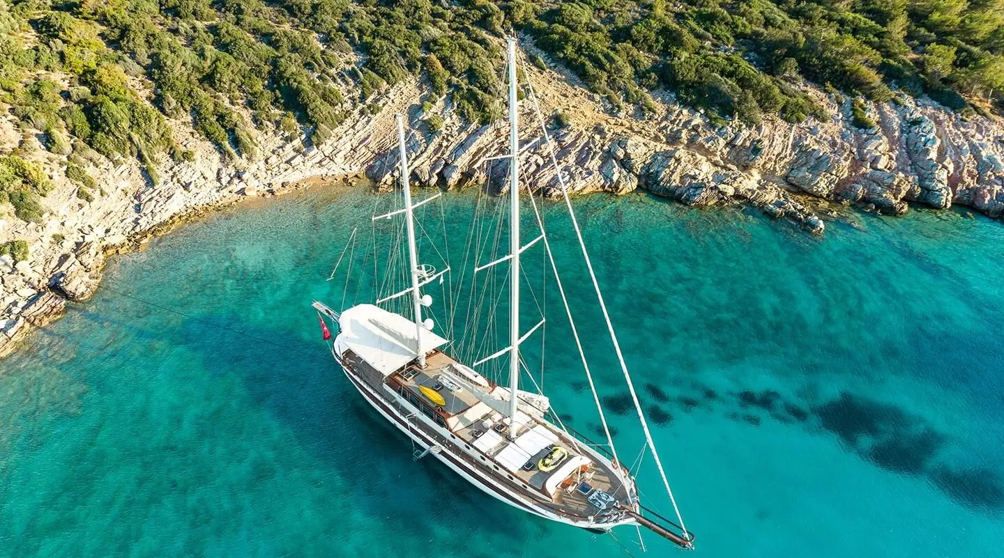 A white sailing yacht anchored in turquoise water near rocky shore at sunset