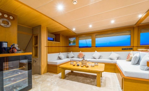Spacious seating area with wood furniture and white cushions, central table with decorative items; blue sea visible through windows outside