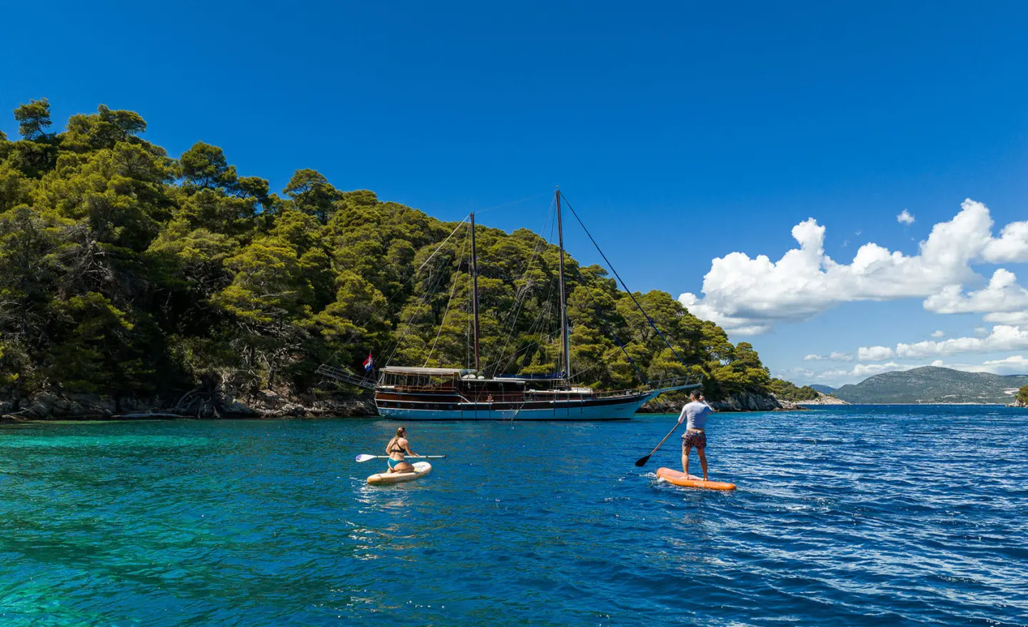 Eleganza yacht anchored in turquoise water with two people swimming near a tree-lined rocky coastline
