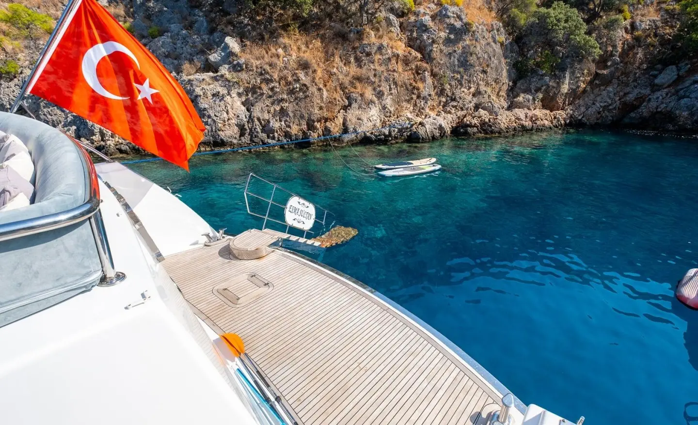 Esma Sultan yacht moored near rocky cliff, sunlit deck and clear blue sea