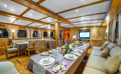 Large wooden interior with ceiling beams, long dining table and chairs, luxury lit dining area on a yacht