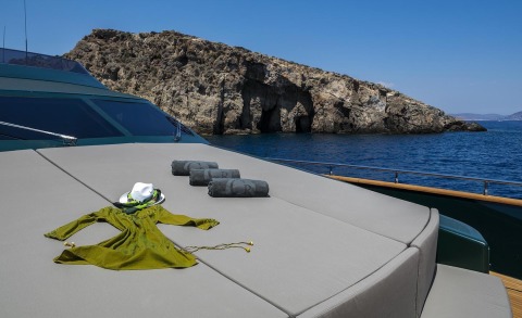 Sunny open sea with the yacht's forward deck, cushions and towels laid out, rocky coastline in the background.