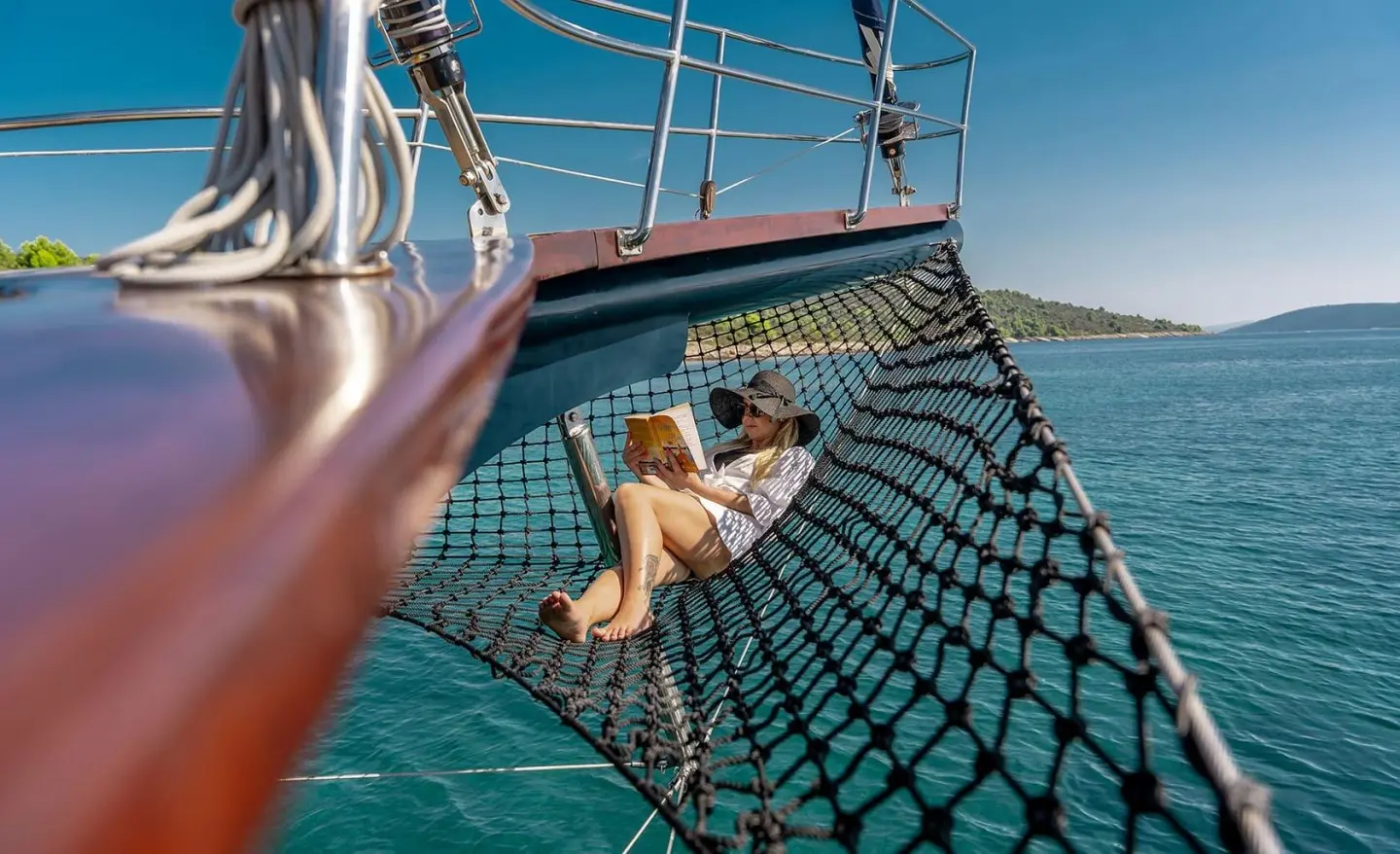 Woman in a net hammock reading a book on a yacht over blue sea at sunset