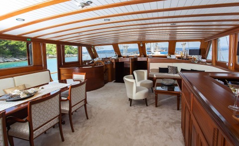 Interior of a wooden yacht lounge with dining table and seating, windows showing water and coastline