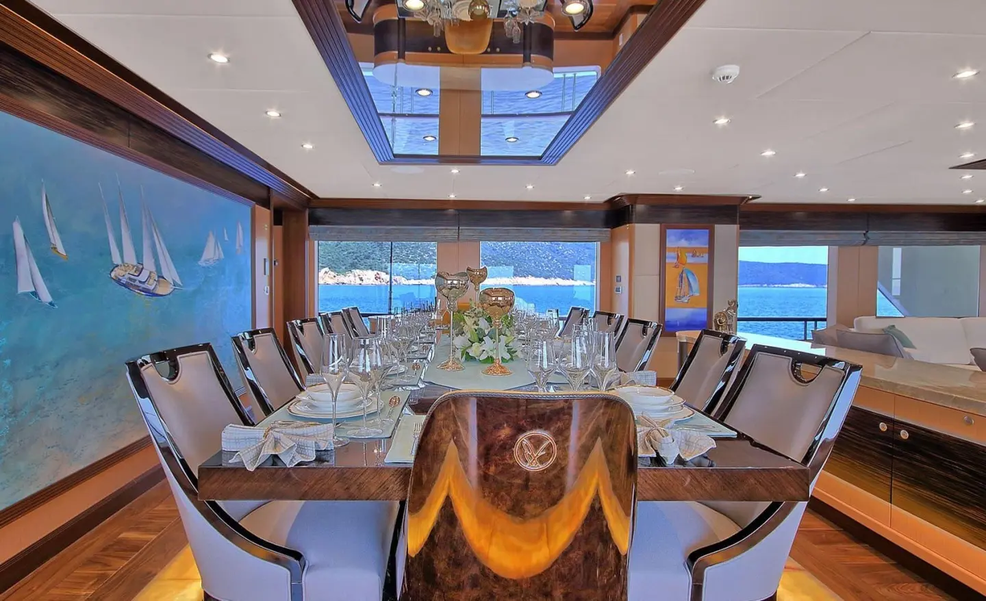 Meira yacht dining area with circular wooden table set for a meal, surrounding chairs, blue walls, and large windows