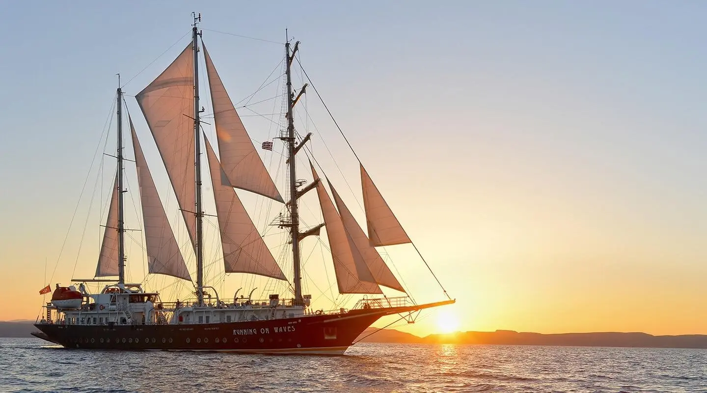 Yacht named Running on Waves sails at sunset on calm sea, red sails prominent