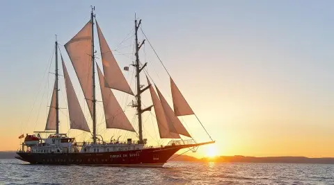 Yacht named Running on Waves sails at sunset on calm sea, red sails prominent