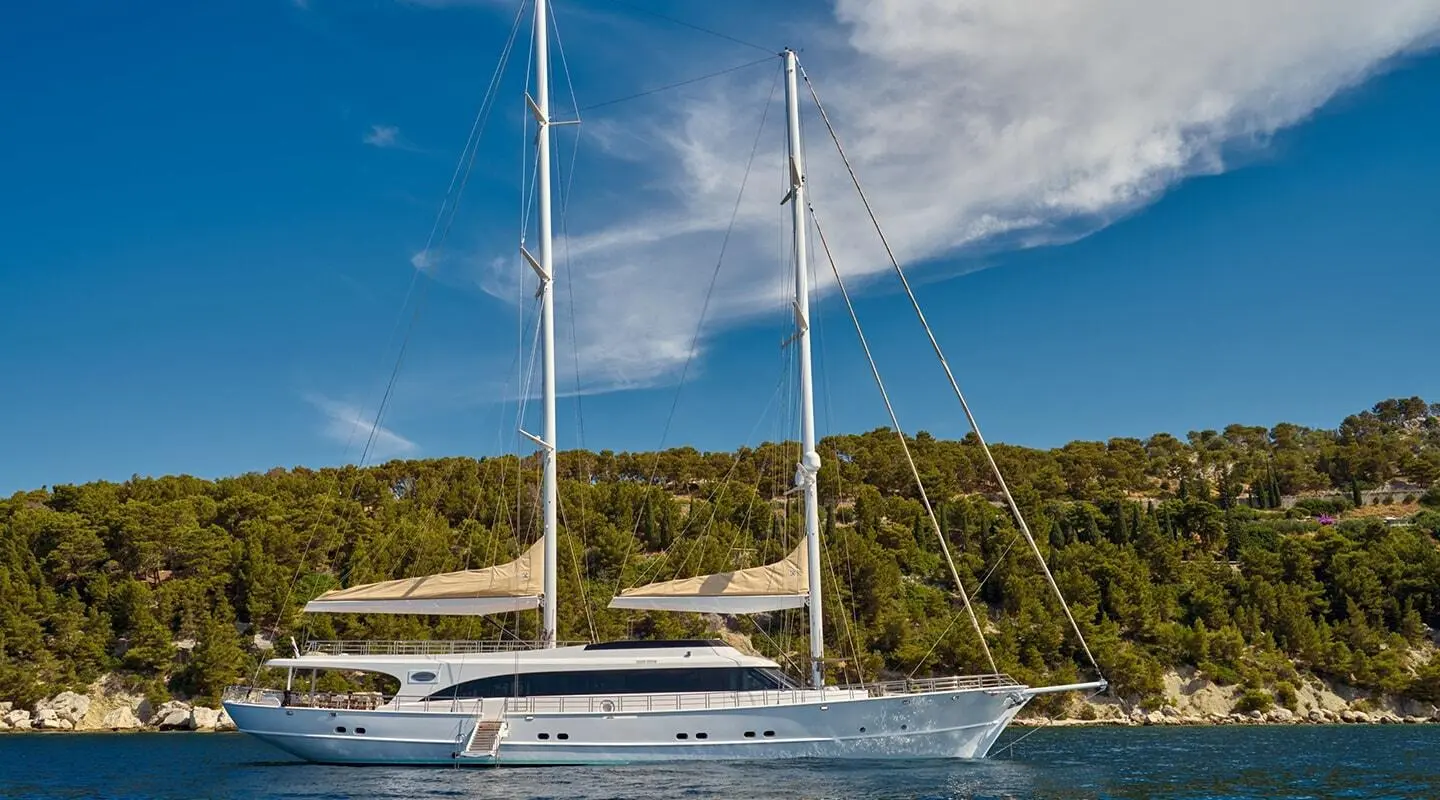 Two-masted yacht anchored by a forested shoreline, calm sea