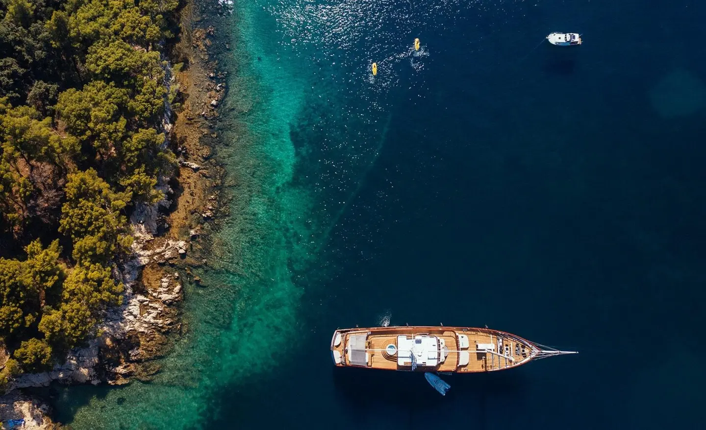 A luxury gulet yacht named Corsario sits anchored near a rocky turquoise shoreline at sunset