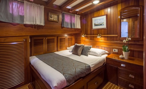 Wood-panelled interior with a large bed, pillows, bedside table and plant decoration inside a yacht cabin