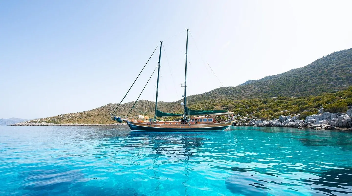 Ros Mare yacht anchored in clear turquoise water near rocky shore at sunset