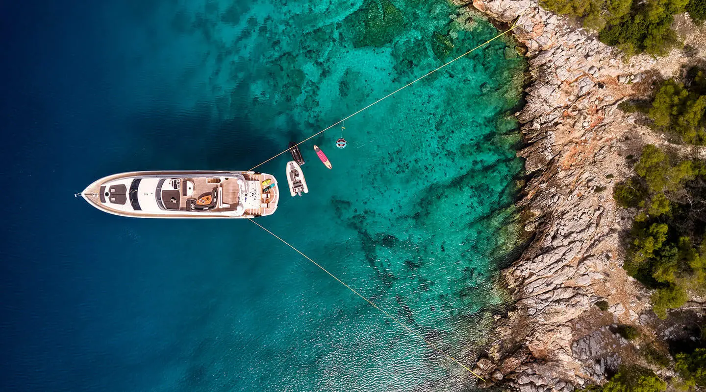 Aerial view of a blue-green sea with a white motor yacht anchored near rocky shoreline