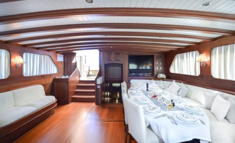 Luxurious yacht interior with a long dining table set with white linens, chairs, wood panels, and windows along the sides
