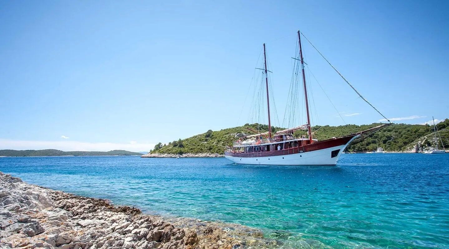 Romanca yacht anchored near rocky shoreline with clear turquoise water and sail masts