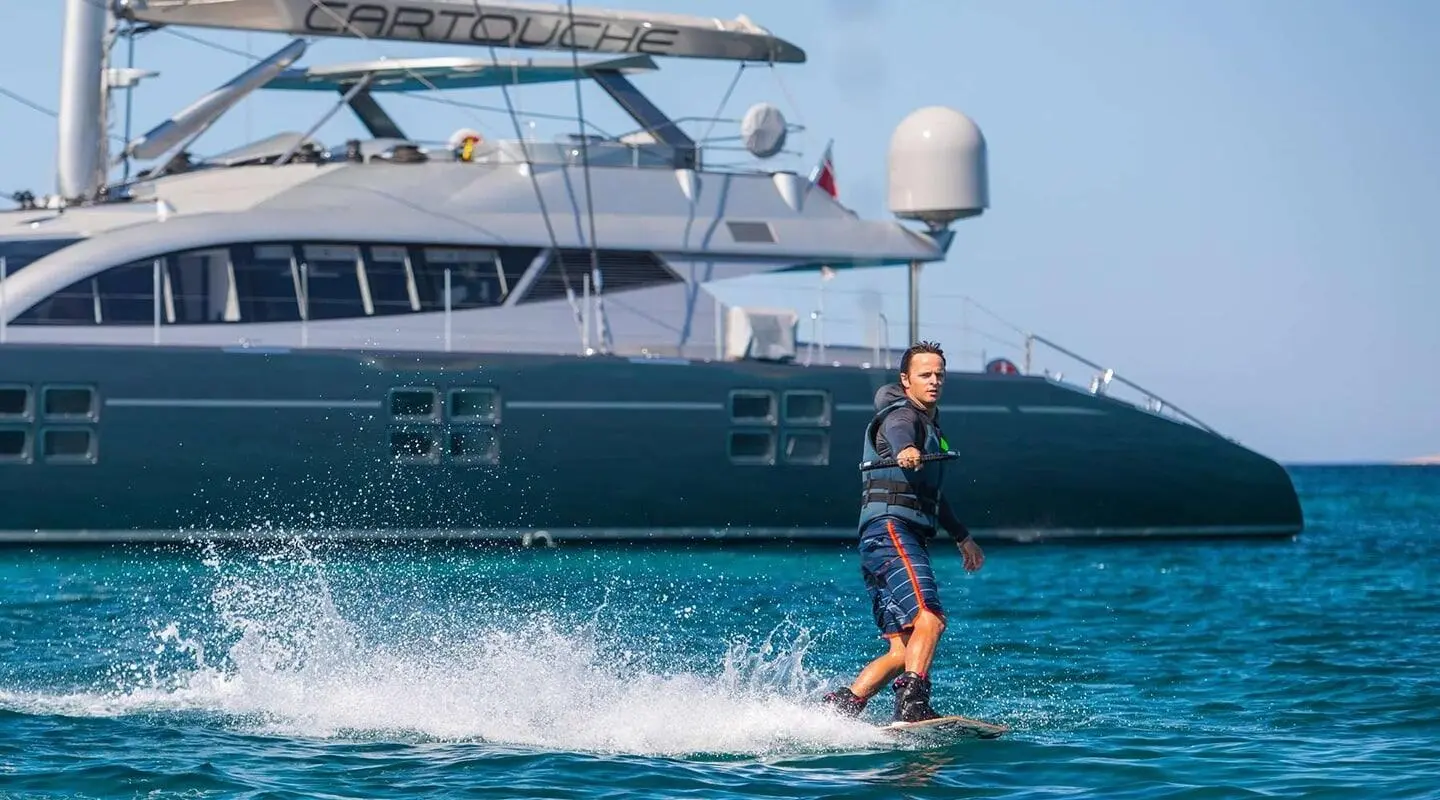 Person wakeboarding near a dark blue luxury yacht Cartouche in clear turquoise water