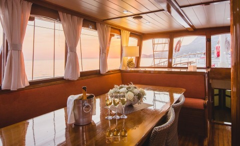 Altair yacht interior: elegant salon with wooden table, champagne, white florals, and sea sunset through windows