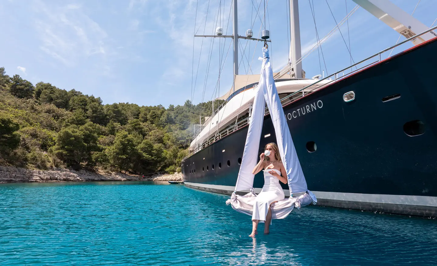 Yacht named Nocturno anchored near rocky coastline at sunset with clear blue water