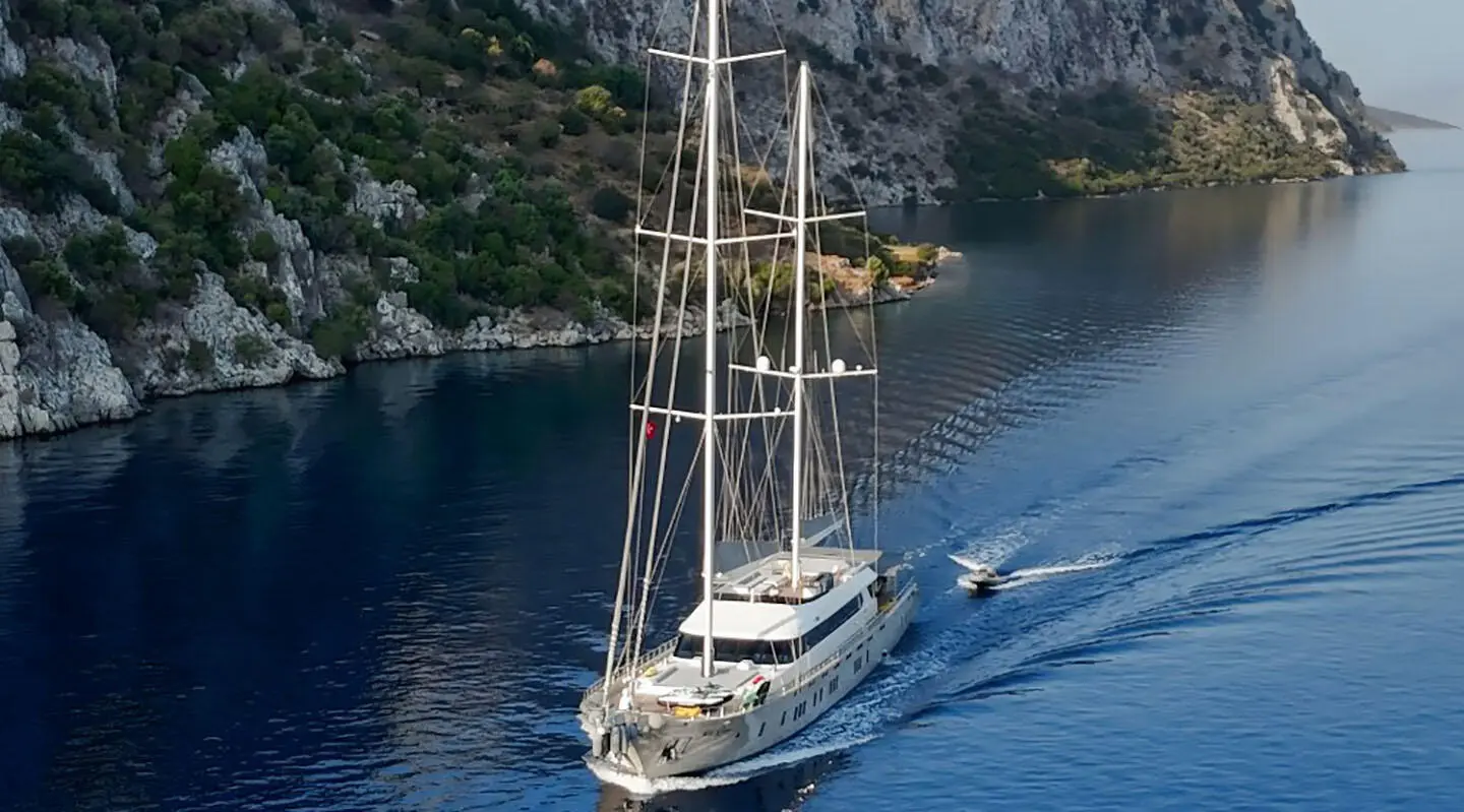 Sailing yacht De Love gliding through blue water near rocky coastline
