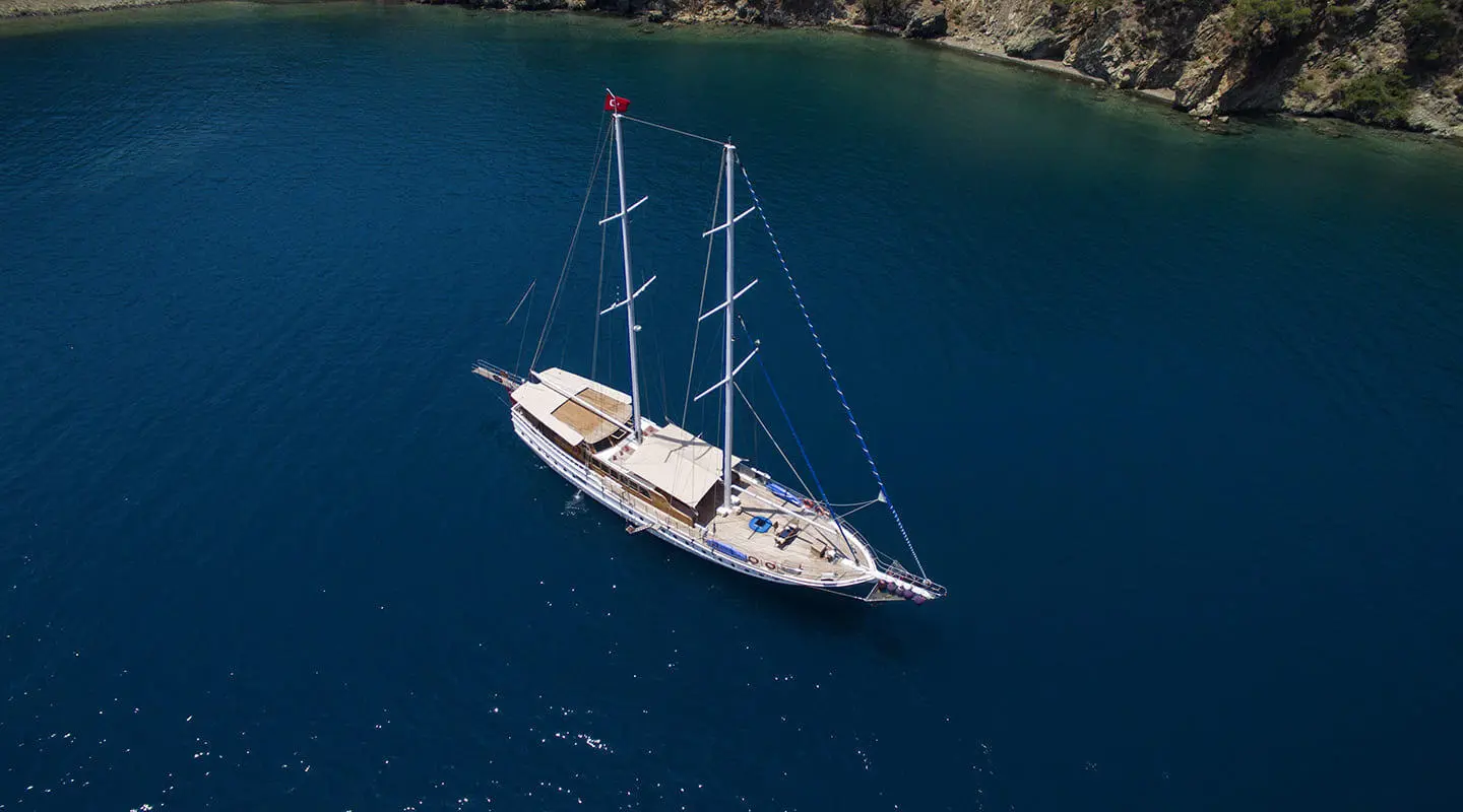 Twin-m mast yacht anchored in blue sea near rocky shore