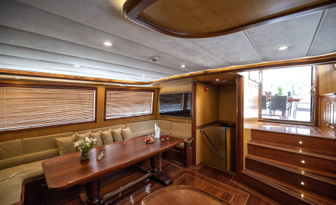 Wooden interior with long dining table and sofa seating; paneled windows on the sides and stairs leading to a deck with outdoor seating visible