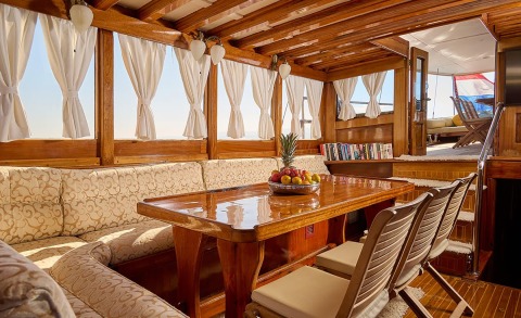 Wooden yacht interior with windows, beige patterned seating, and a polished table with a fruit bowl