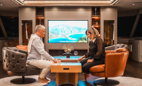 Two people seated in a yacht salon, facing each other at a wooden table with drinks