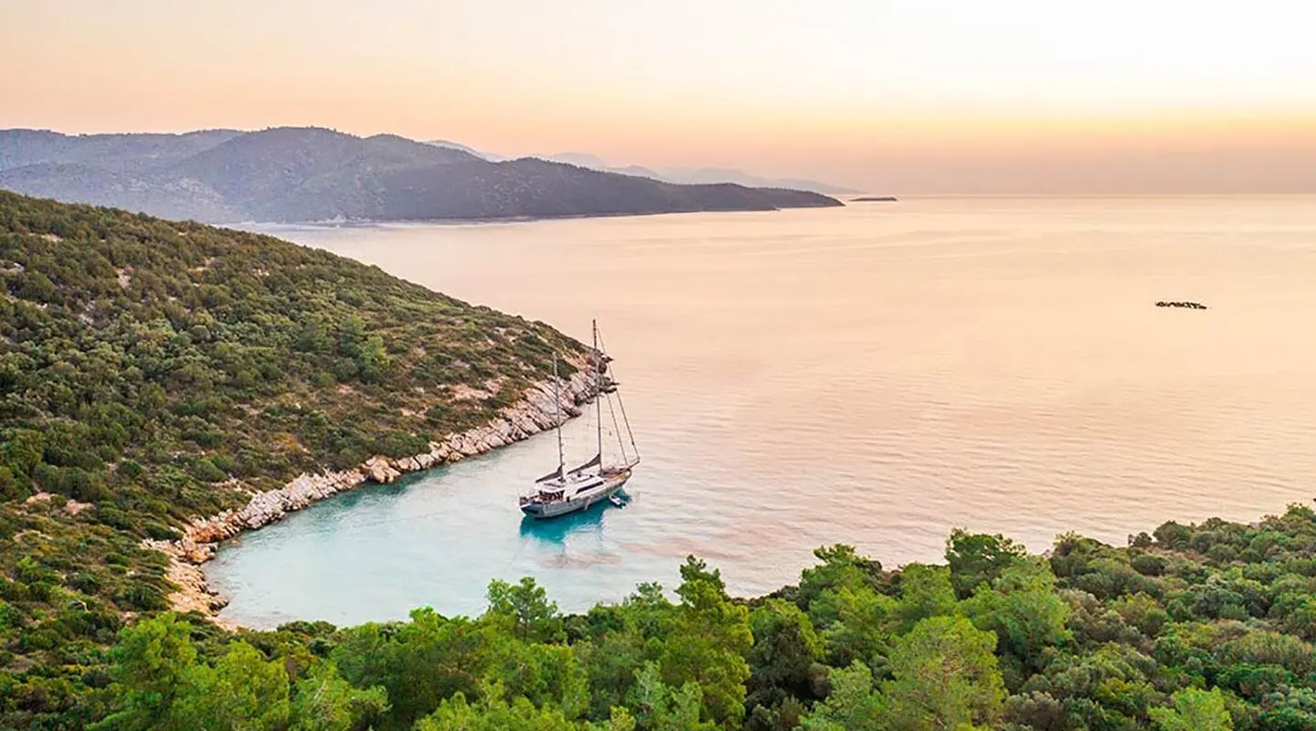 Virtuoso yacht anchored along a rocky coastline at sunset, calm water